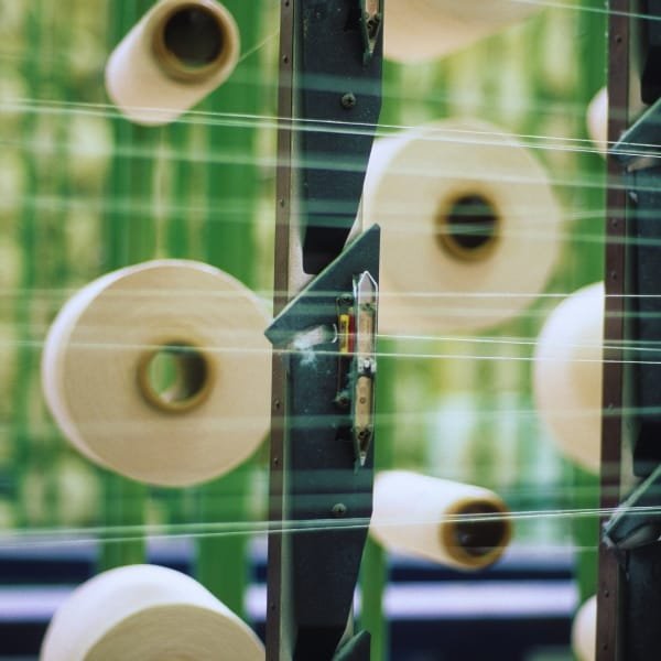 A textile manufacturing setup with multiple large spools of beige paper or fabric rolls mounted on a metal frame. Spools are arranged in rows, with thin horizontal lines (threads or strings) crossing the scene. The background shows a blurred green environment.
