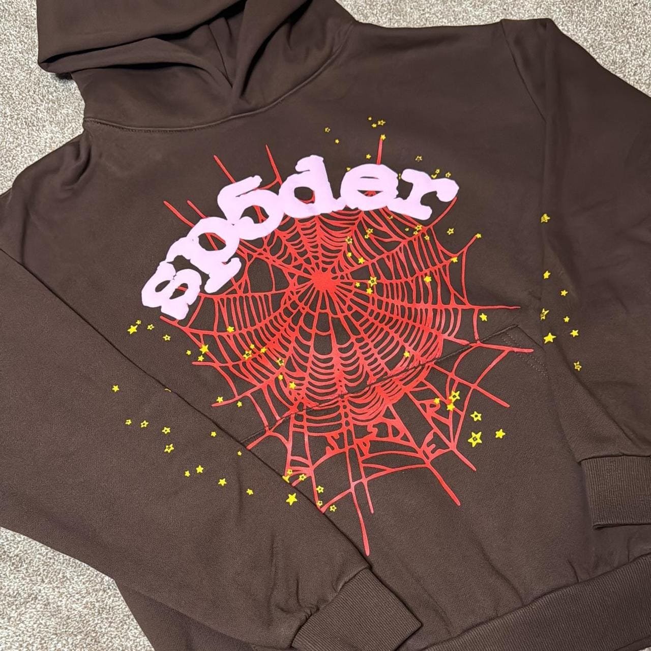: A brown hoodie featuring a large red spiderweb graphic with the word “spider” in chunky white/purple lettering across the top and small yellow stars scattered around the web.