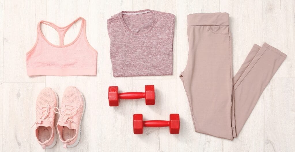 Flat lay of a pastel pink workout ensemble: sports bra, heather pink tee, light pink leggings, pink sneakers, and two red dumbbells on a light wooden floor.