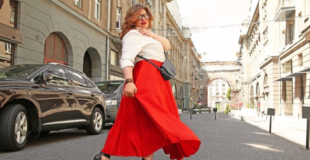 Plus size woman wearing a white shirt, red flowing skort, and heels, walking on a city street under bright sunlight