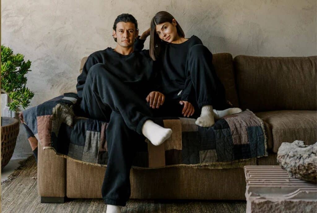 "A designer sweat suit made from organic cotton, in earth-toned colors, displayed on a mannequin in a bright, minimalist showroom with a USA flag graphic in the background to signify American sourcing."