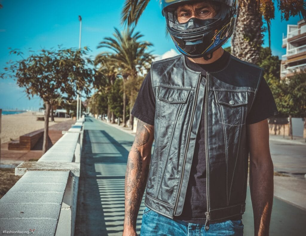 A person wearing a black leather vest and helmet, standing on a scenic road by the beach, showcasing stylish and durable motorcycle gear.