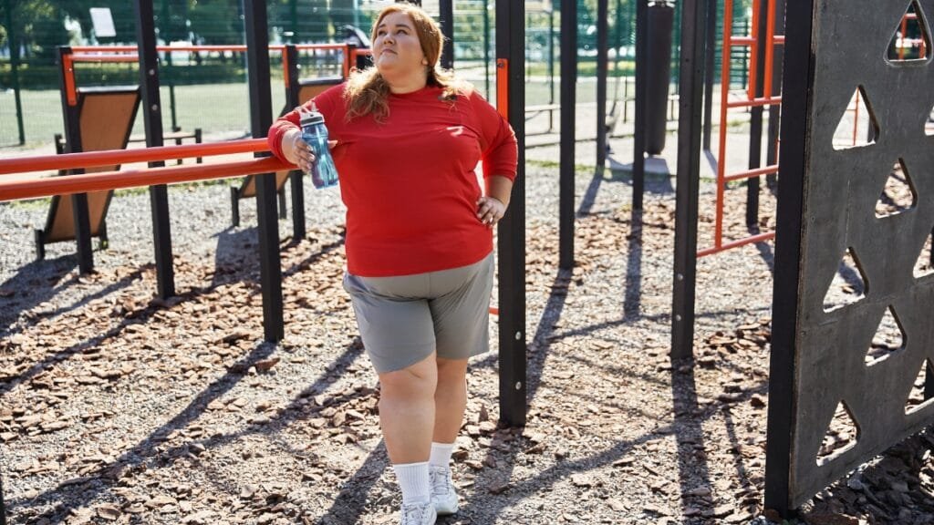 Plus size woman wearing a red shirt and gray walking shorts set, standing outdoors at a playground with a water bottle