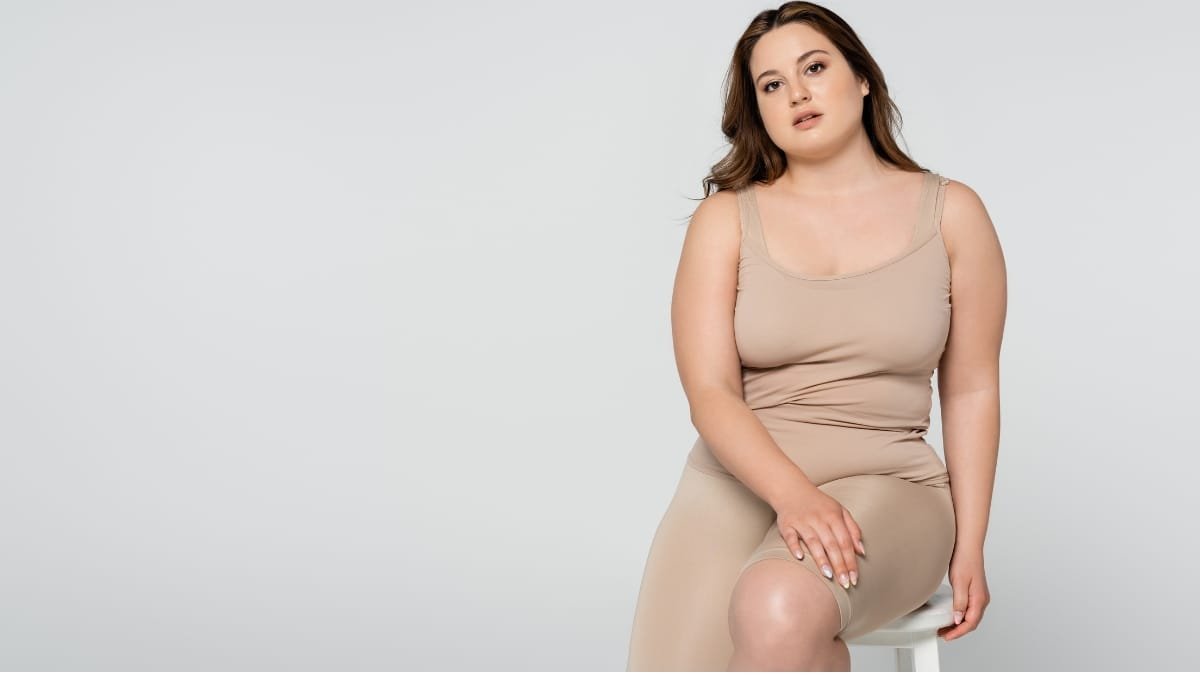 Woman confidently wearing a tan plus size dress, sitting on a chair with a calm expression