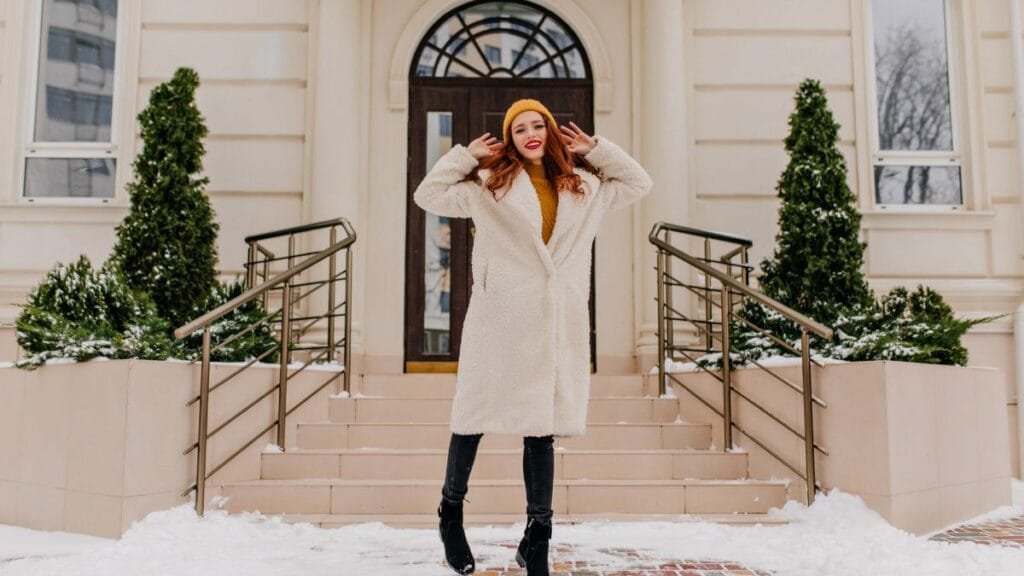 A woman wearing a cozy white teddy coat, yellow beanie, and knee-high boots, standing on snowy steps in front of a building, smiling and enjoying the winter day.