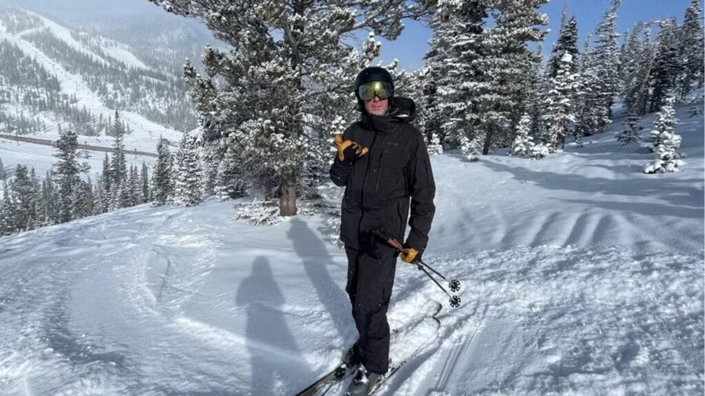 "Skier wearing a black mens ski jacket, standing in snowy mountains with a scenic winter backdrop."