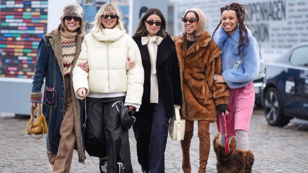 A group of five women standing together outdoors, dressed in stylish winter outfits. Each woman is wearing a different fashionable coat, with accessories like sunglasses, scarves, and bags. They are smiling and posing in front of a colorful backdrop, showcasing their winter fashion looks.