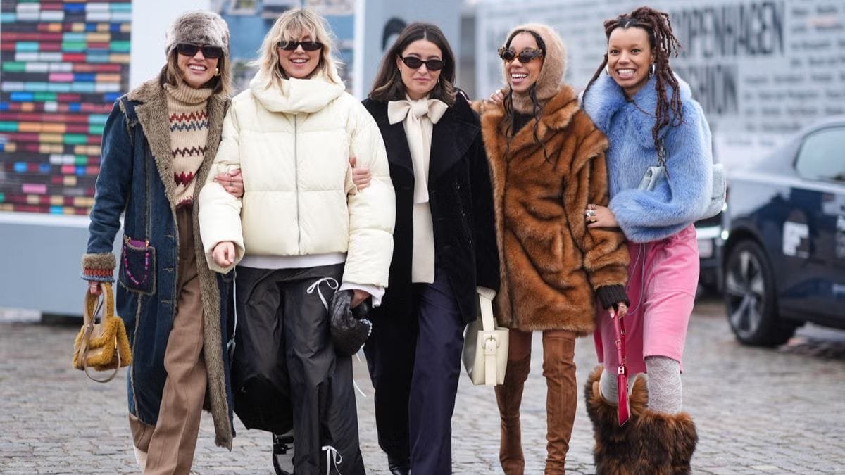 A group of five women standing together outdoors, dressed in stylish winter outfits. Each woman is wearing a different fashionable coat, with accessories like sunglasses, scarves, and bags. They are smiling and posing in front of a colorful backdrop, showcasing their winter fashion looks.