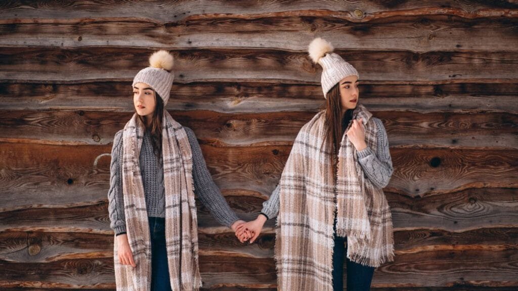 Two women dressed in cozy knit hats and plaid scarves, holding hands while standing against a wooden wall. Both are wearing gray sweaters and denim jeans, showcasing cute winter outfits with plaid and checks