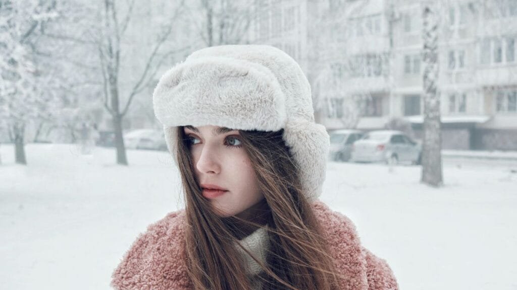 Woman wearing a fluffy winter hat and pink coat in a snowy setting.