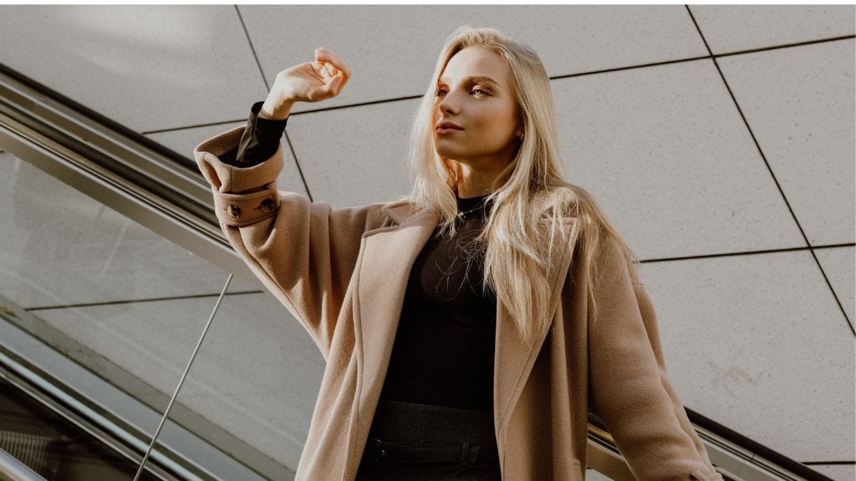 “Woman wearing a stylish beige coat standing near an escalator in modern architecture.”