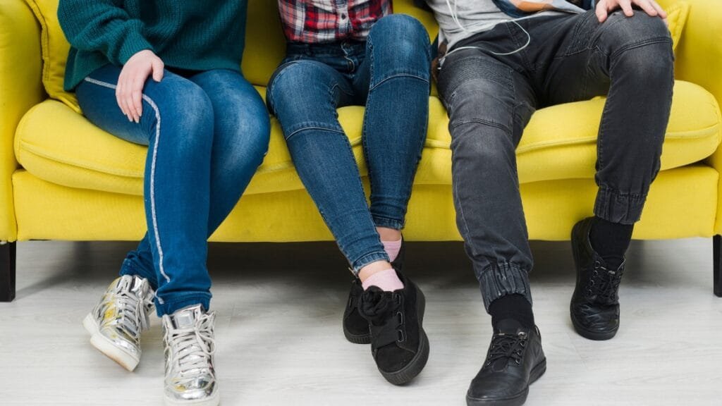 Three people sitting on a yellow sofa showing only their legs and footwear, wearing casual jeans and sneakers.
