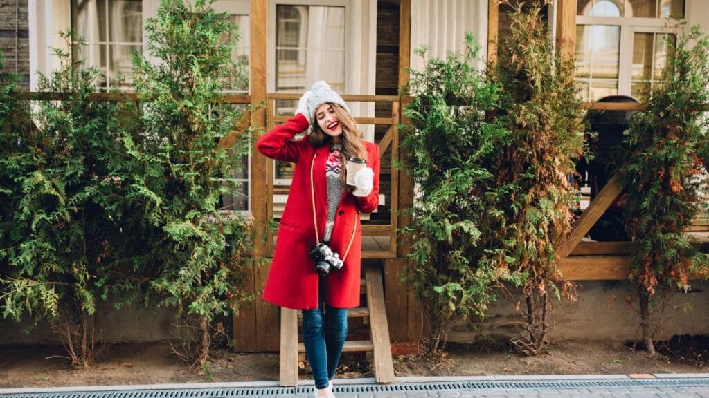 A smiling woman wearing a red coat, gray sweater, white hat, and gloves, holding a coffee cup and a camera, standing outdoors in front of a wooden house with green shrubs.