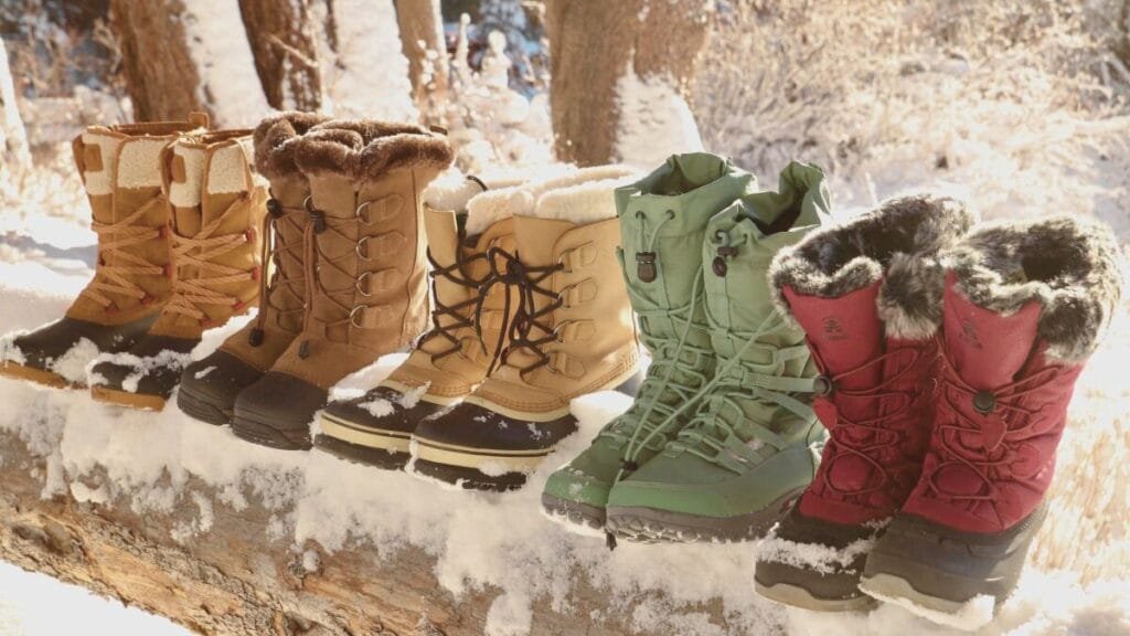 A row of winter boots in various colors—brown, tan, green, and red—lined up on a snow-covered log in a snowy outdoor setting.