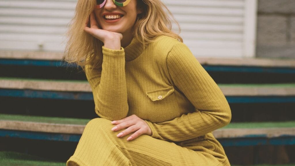 Smiling woman with blonde hair wearing colorful sunglasses and a yellow ribbed sweater, sitting on outdoor stairs.