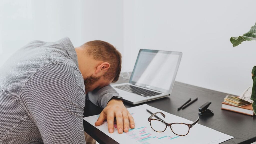 A person sitting at a desk, feeling overwhelmed, with their head resting on the table next to a laptop and papers.