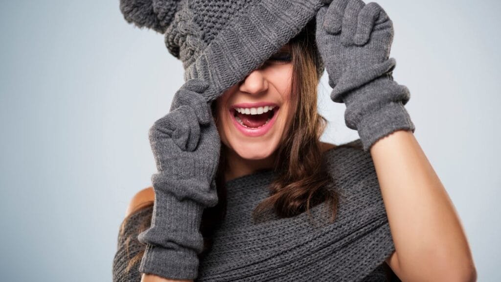 A woman smiling while playfully covering her eyes with a gray knitted hat and gloves, both made of cozy, soft fabric, against a light background.