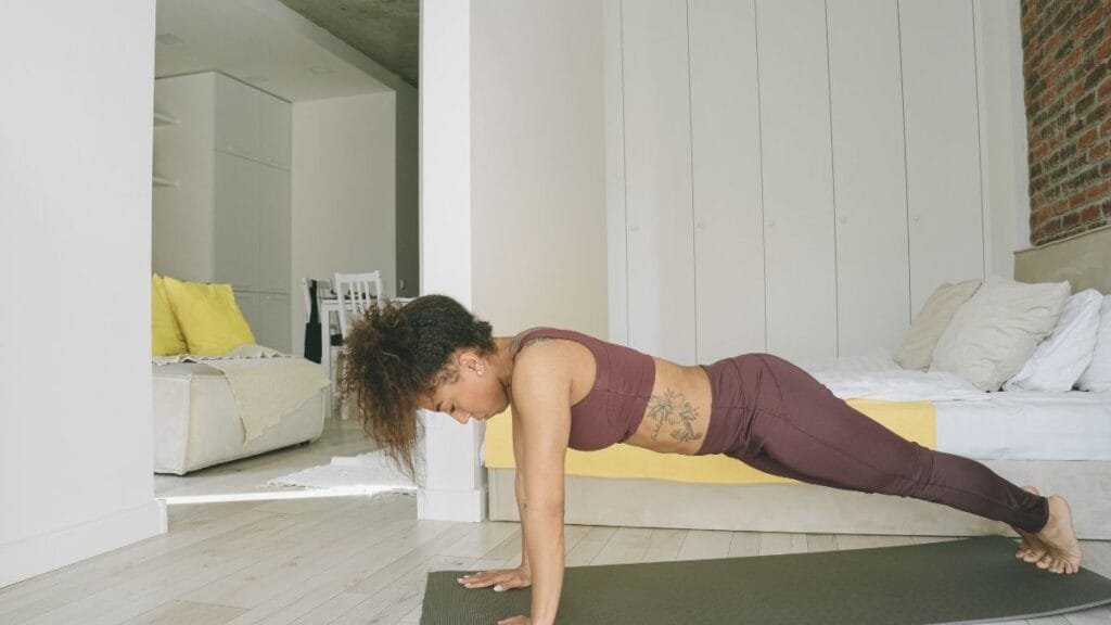  Woman in maroon workout attire performing a plank exercise on a yoga mat in a bright, minimalist room.