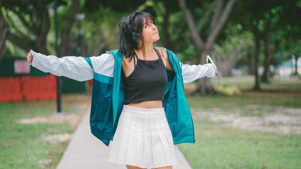 Woman enjoying music outdoors in a tennis skirt, black crop top, and a teal jacket.