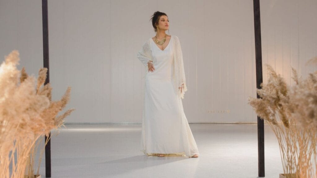 A woman wearing a formal white midi dress with sleeves, accessorized with a layered necklace, standing gracefully in a minimalistic indoor setting with decorative dried plants in the foreground.