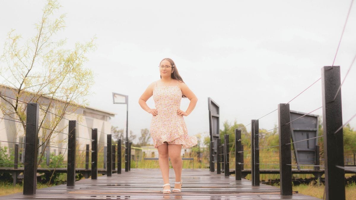 A confident woman stands on a wooden bridge, wearing a floral dress and sandals, with trees and buildings in the background.