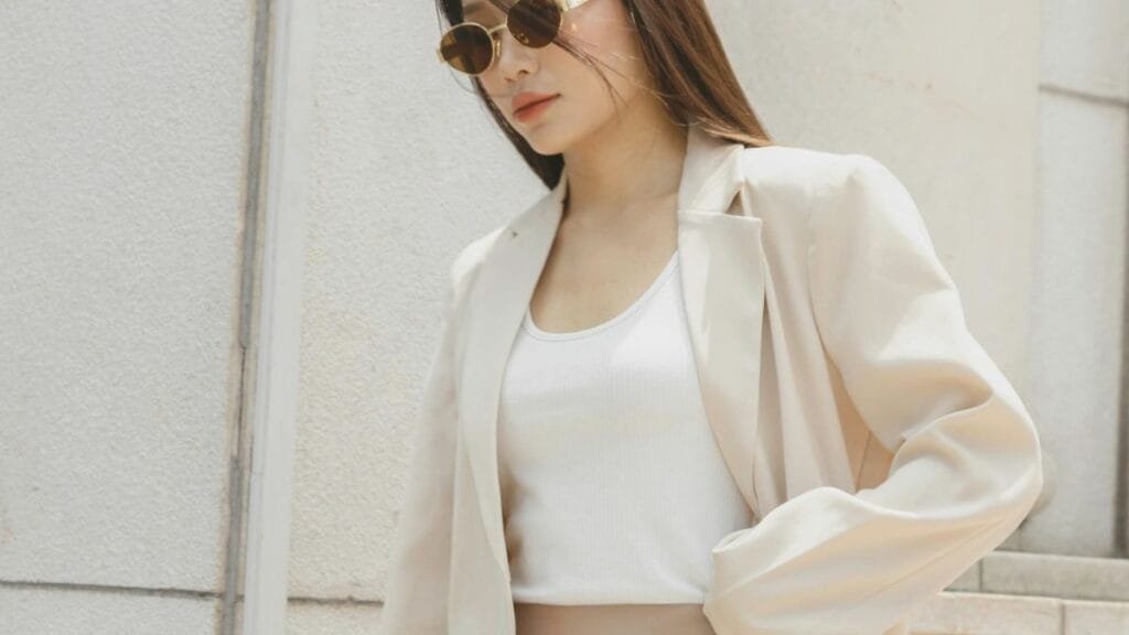 A woman wearing a white tank top, beige blazer, and sunglasses, walking in an outdoor setting with a neutral-colored background.