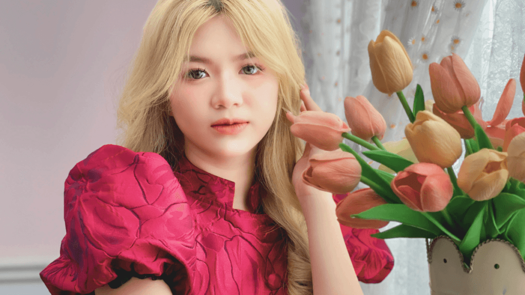 Young woman with blonde hair wearing a pink floral dress, posing next to a bouquet of peach-colored tulips with a soft expression