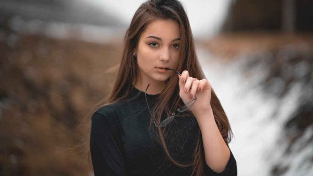 Young woman with long brown hair, wearing a black shirt, holding sunglasses in her mouth, looking at the camera with a serious expression.