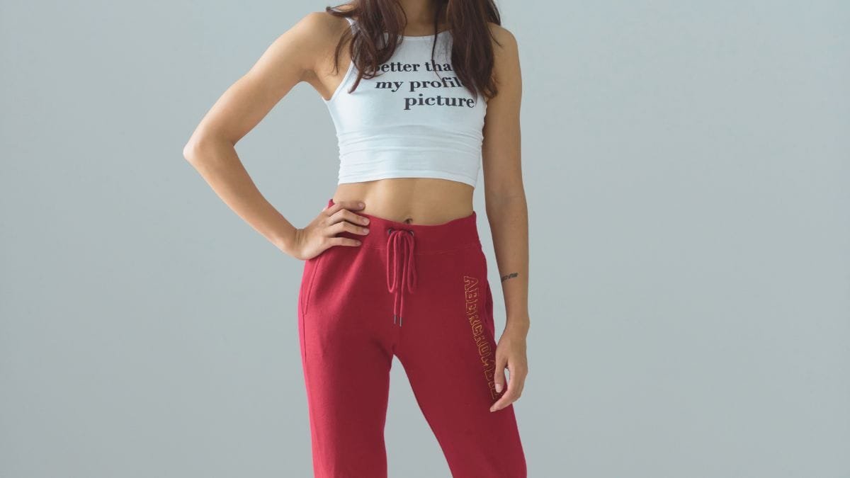 A woman wearing a white cropped top with the text "Better than my profile picture" and red sweatpants with a drawstring.