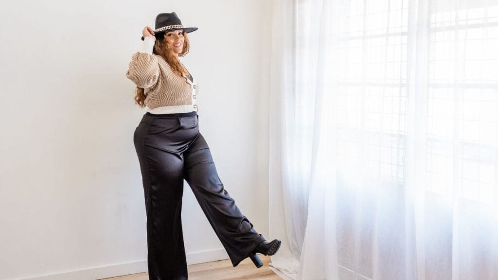  A woman wearing a beige cardigan, black trousers, and a black hat, striking a playful pose in front of a window with natural light.