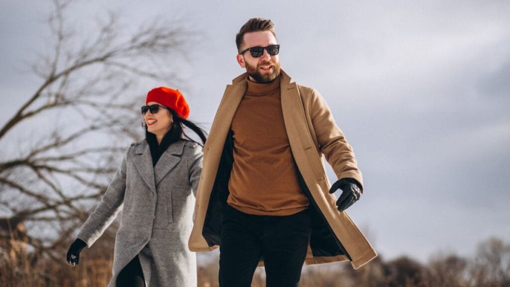 A couple walking in the snow, both dressed in stylish winter coats, with the woman wearing a red beret and the man a beige coat, looking fashionable.