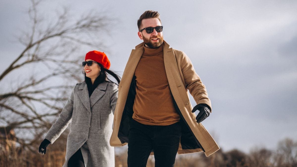A couple walking in the snow, both dressed in stylish winter coats, with the woman wearing a red beret and the man a beige coat, looking fashionable.