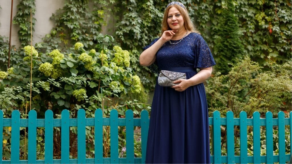 Plus-size woman in a navy blue dress with lace details, standing in a garden with a silver clutch and a tiara.