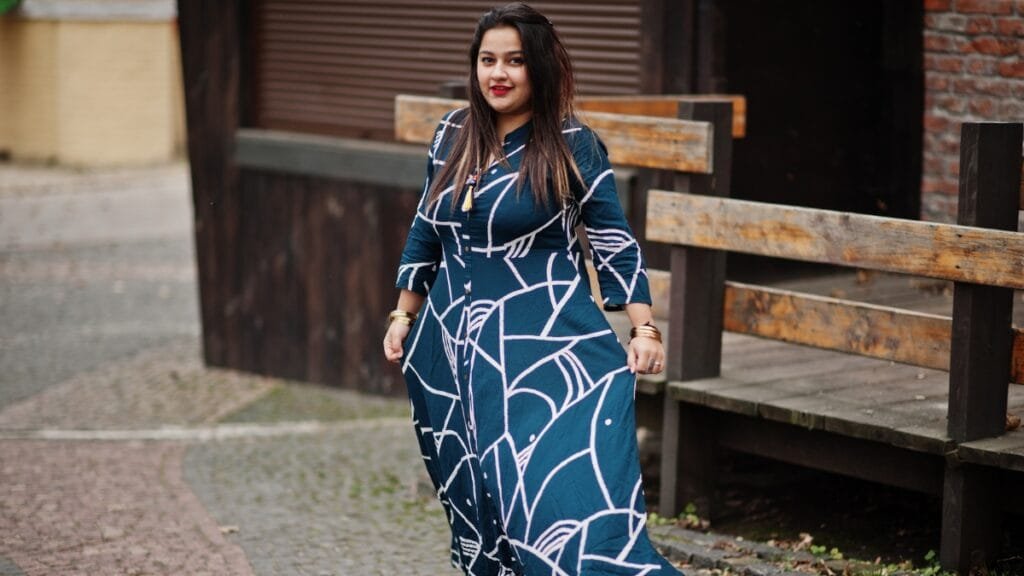 Plus-size woman in a blue dress with abstract white patterns, smiling while walking outdoors.