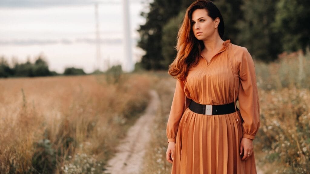 A woman in a flowing brown plus size dress standing in a field with a serene expression, showcasing the dress's flattering fit