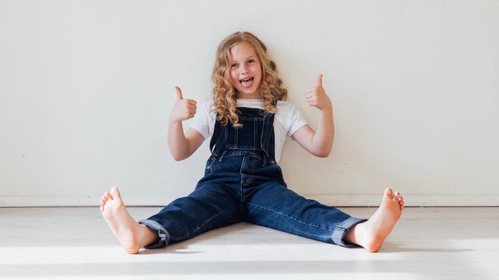 A 9-year-old girl wearing denim overalls, a t-shirt, and smiling while sitting on the floor