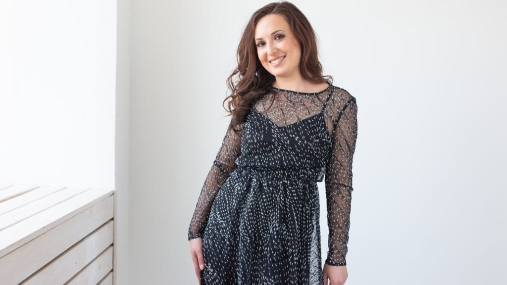 Curvy woman in a black sheer long-sleeve cocktail dress, size 14, standing near a window with natural light