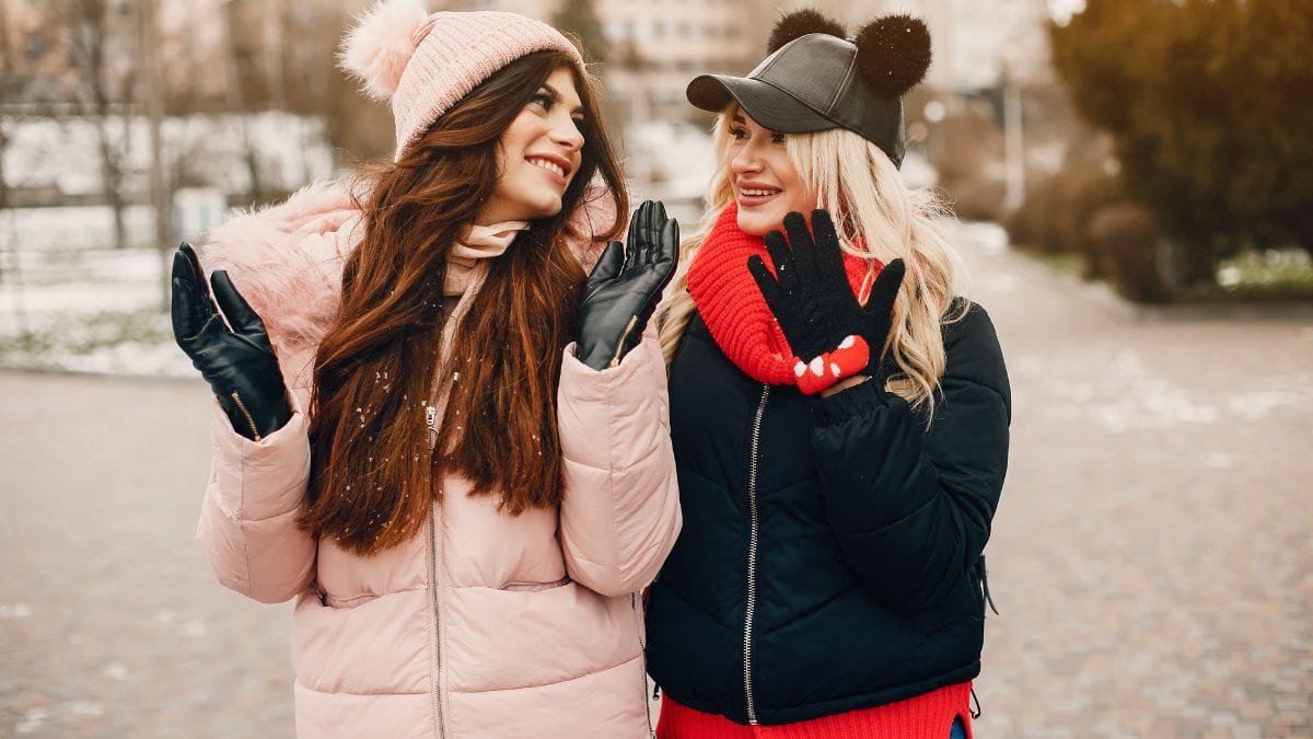 Two women enjoying the winter weather, dressed in stylish coats, hats, and gloves, smiling and waving at each other.