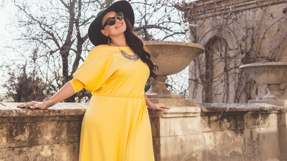 A woman in a yellow dress posing outdoors near a stone structure, smiling with a hat and stylish accessories