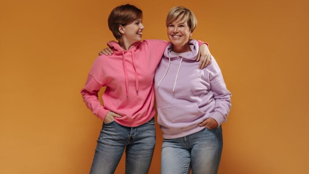 Two women wearing plus size sweaters, one in a pink hoodie and the other in a lavender hoodie