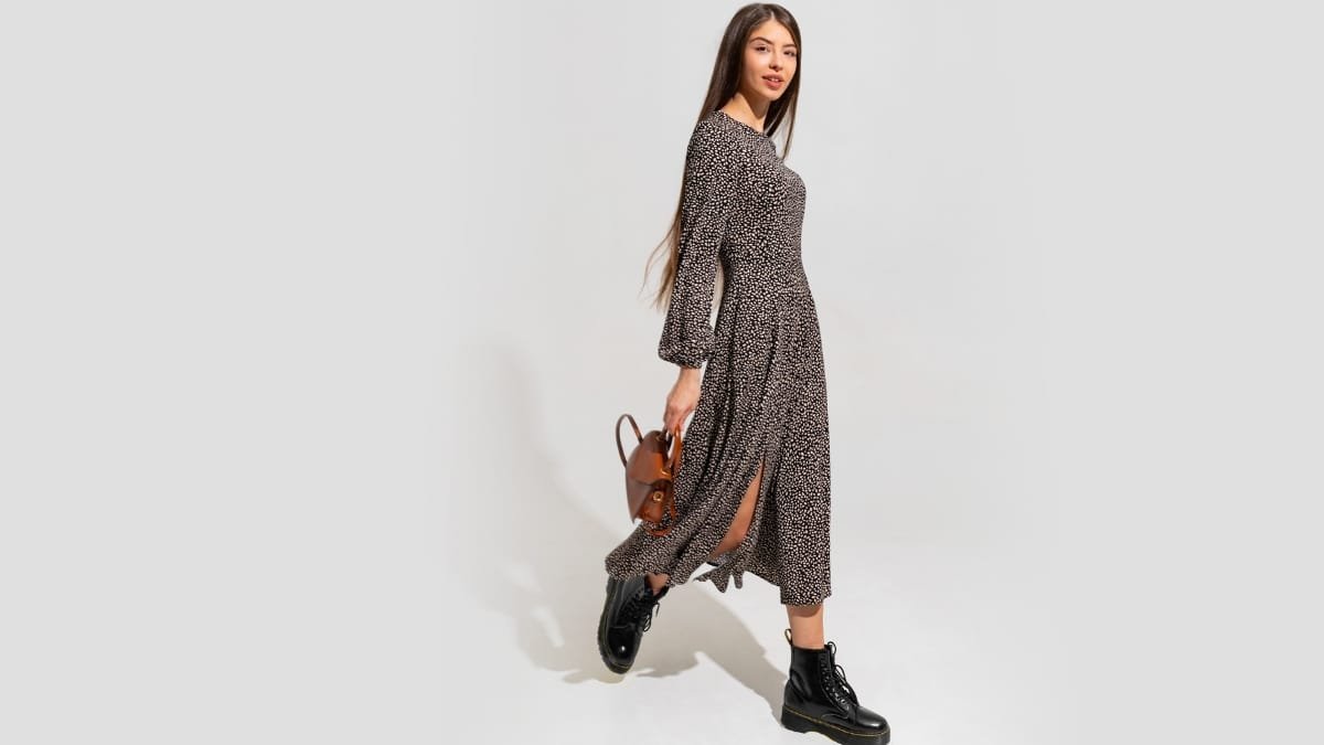 Model wearing a long sleeve midi dress with black boots and a brown handbag, walking in a studio setting