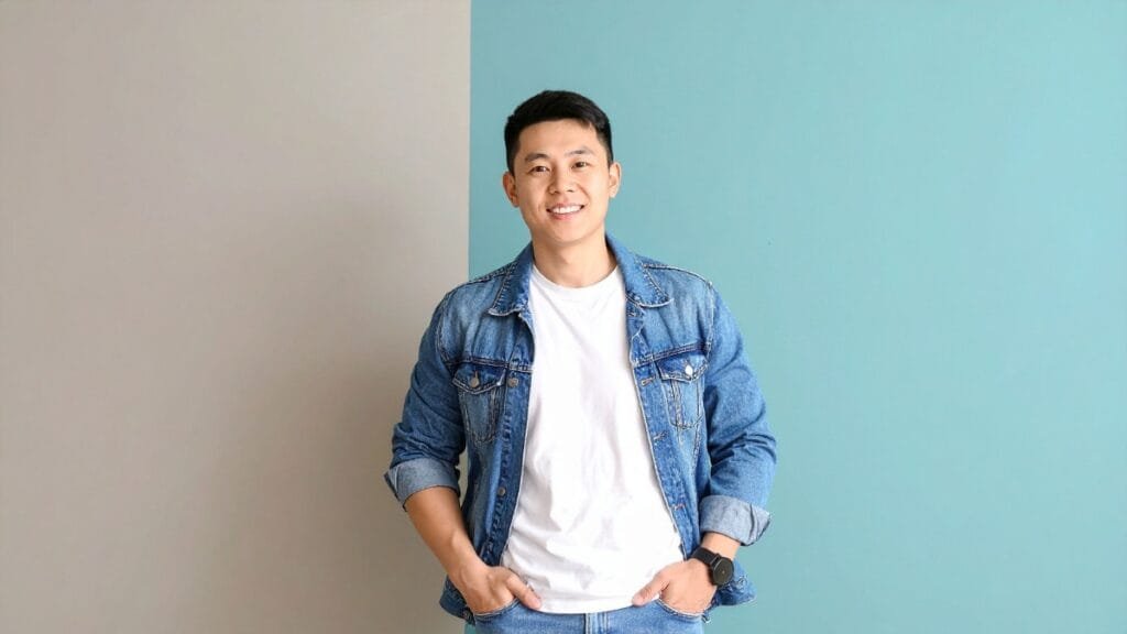 A person confidently wearing a tall denim jacket with a simple white t-shirt, smiling in front of a pastel-colored wall