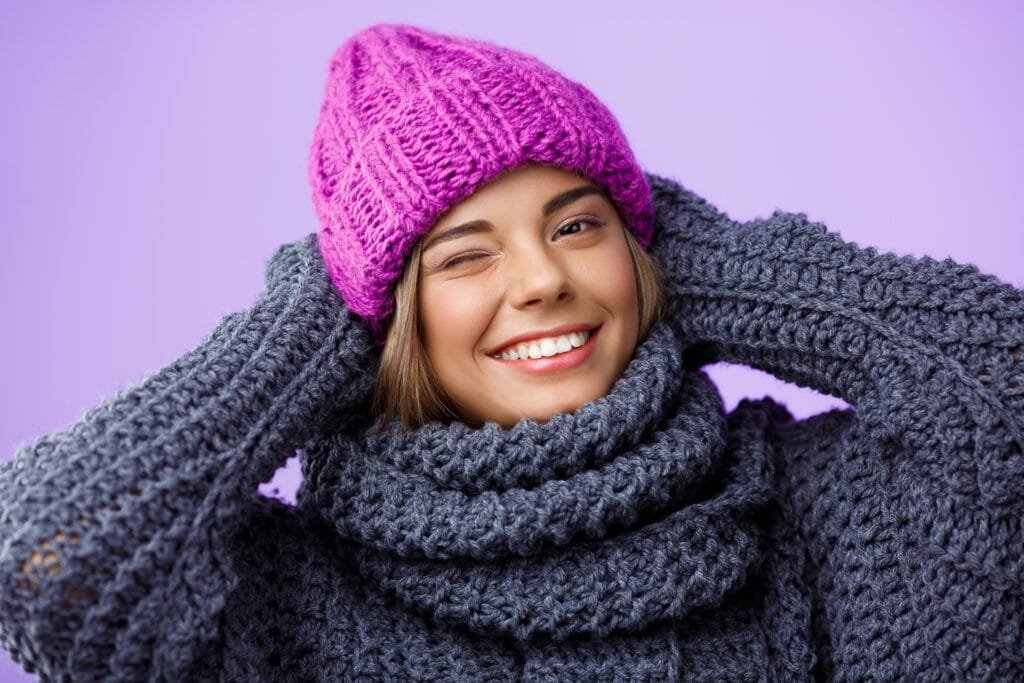 A young woman wearing a cozy purple knitted hat and a gray chunky knitted sweater and scarf, smiling and winking at the camera. The background is soft violet, complementing her cheerful expression.
