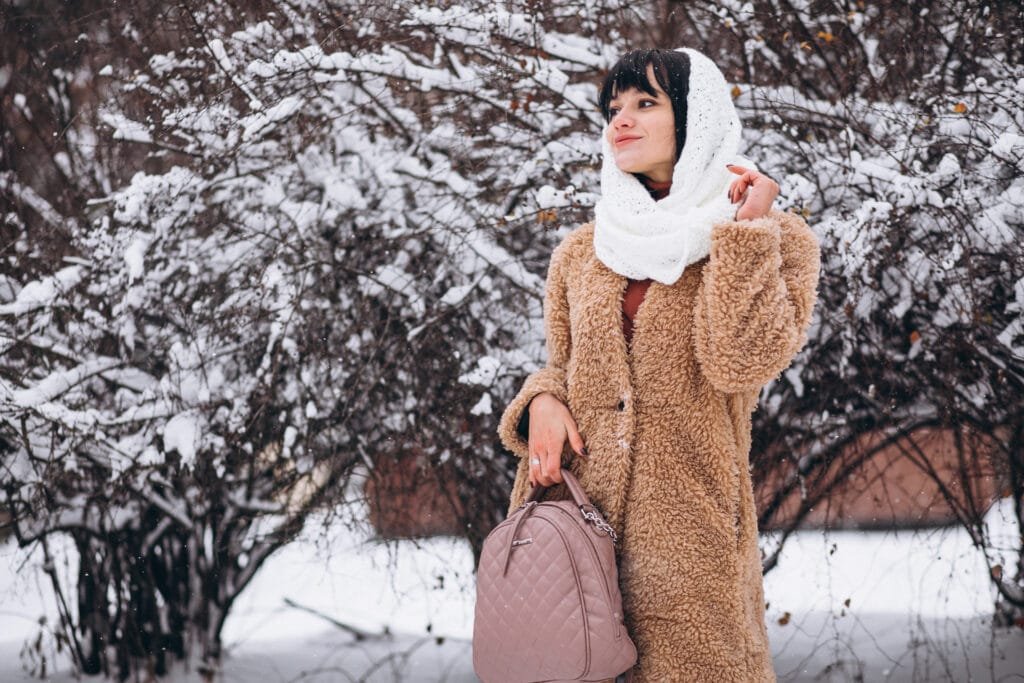 A young woman in a fluffy beige coat and white scarf, holding a pink backpack, smiling in the snow.