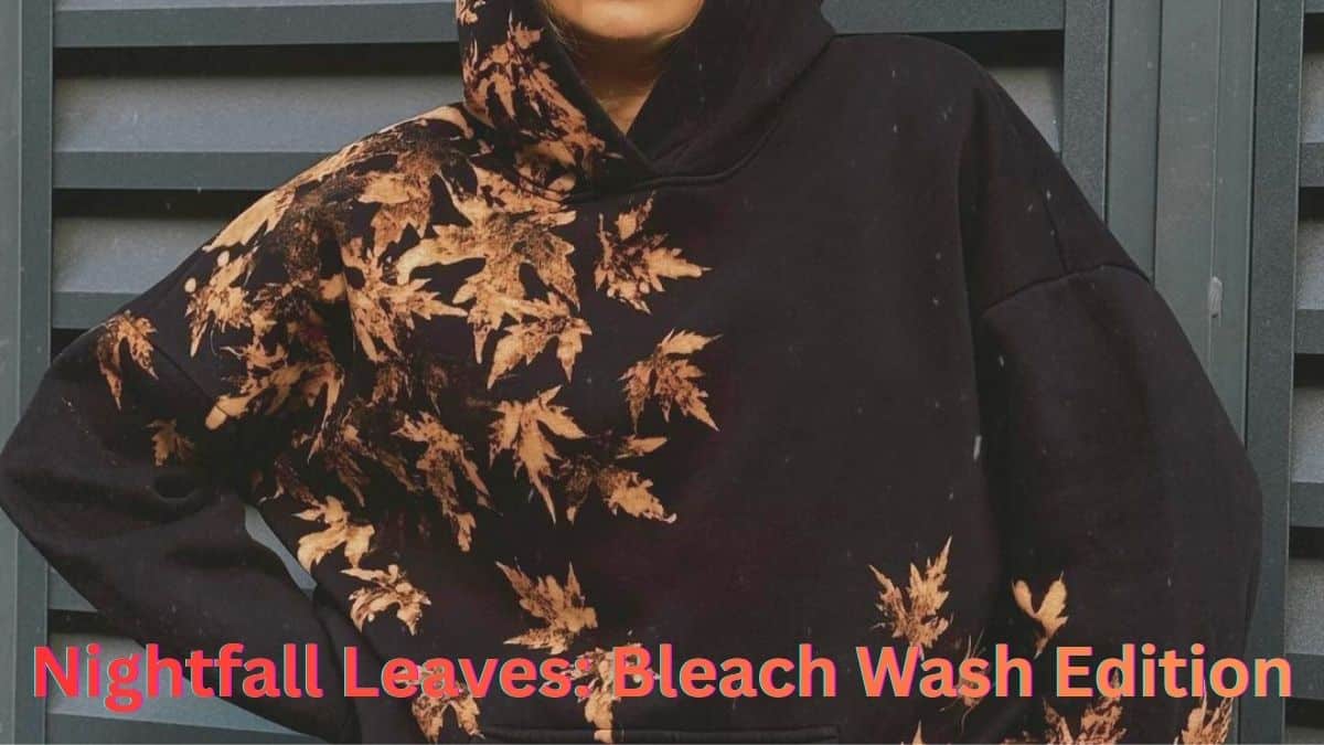 Person wearing a black hoodie with orange bleach-printed maple leaf pattern, standing against a blue-gray slatted wall. The hood is up and hands rest on hips.