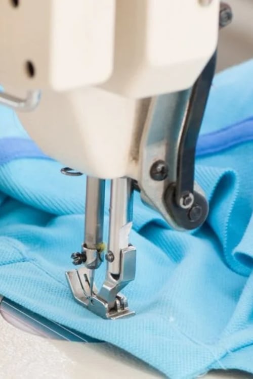Close-up of a sewing machine needle and presser foot stitching turquoise fabric.