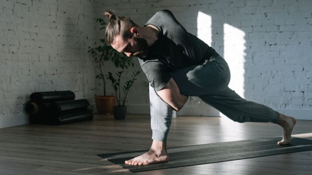 "Man practicing yoga in a bright, spacious room with wooden floors and yoga mats stacked in the background. He is wearing a dark shirt and light gray pants, performing a stretching pose on the yoga mat." This alt text describes the image in a clear and concise manner while being SEO-friendly and accessible. Here’s a suggested alt text for the image you provided: **"Man practicing yoga in a bright, spacious room with wooden floors and yoga mats stacked in the background. He is wearing a dark shirt and light gray pants, performing a stretching pose on the yoga mat."** This alt text describes the image in a clear and concise manner while being SEO-friendly and accessible.