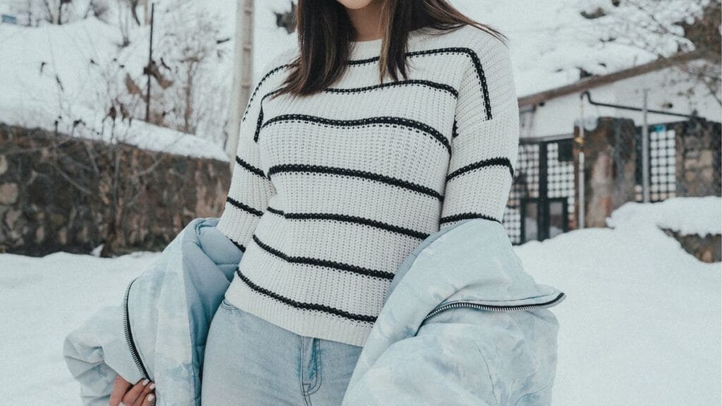 A woman wearing a white striped sweater and light blue jacket standing outside in the snow.