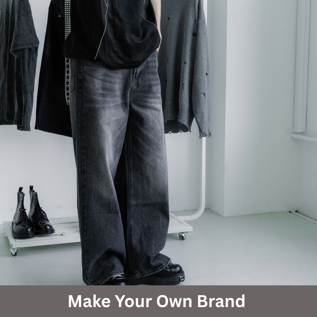 A person stands in a studio wearing oversized black jeans and black shoes, with clothing racks and boots in the background. The bottom overlay reads “Make Your Own Brand.”