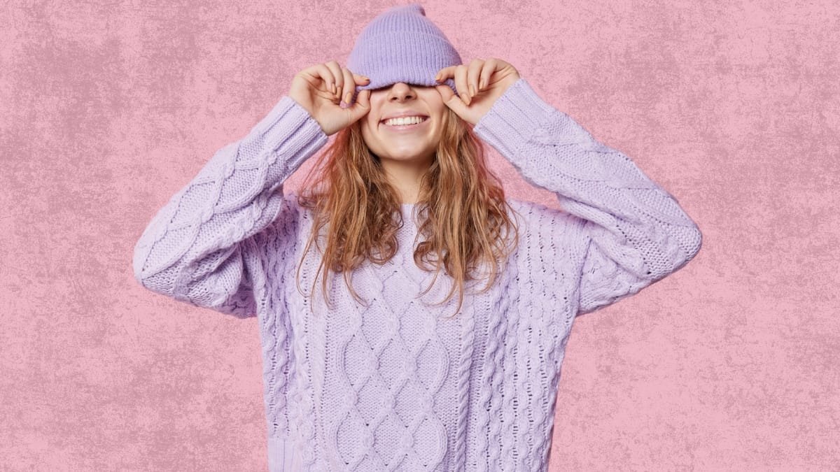 Woman playfully hiding behind her wool hat, wearing a soft lilac wool sweater